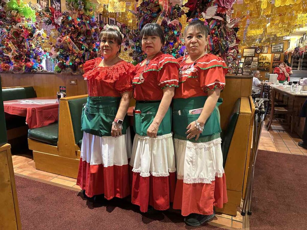 Three female servers in red blouses, green aprons, and white skirts standing together inside Mi Tierra Café surrounded by colourful festive décor