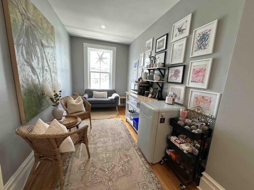 Narrow common area at The Ivy House Inn in Lunenburg, featuring wicker chairs, a small loveseat by the window, a patterned runner, a mini fridge, tea and coffee station, and a gallery wall displaying children’s artwork by Ivy.