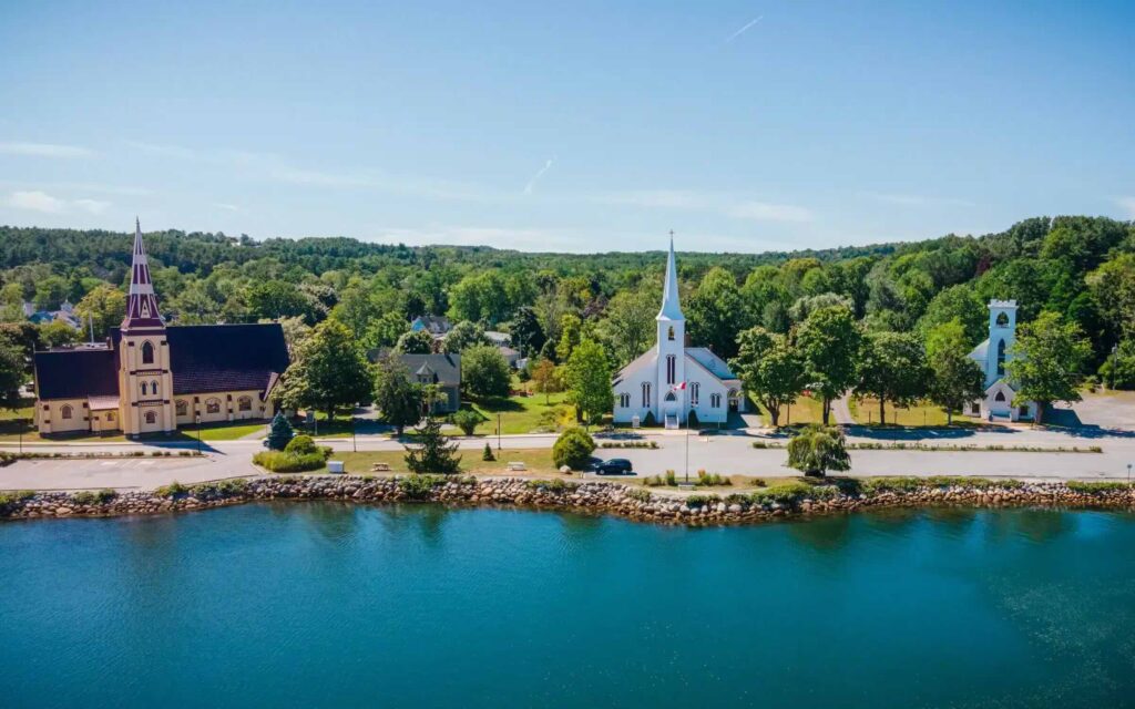 Aerial view of Mahone Bay’s waterfront showing the town’s three iconic churches lined up beside calm blue water, with leafy green trees and small homes stretching into the background.