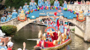 A brightly decorated Texas Cavaliers river barge glides along the San Antonio River beneath a rainbow-fringed bridge, surrounded by participants in light blue uniforms during Fiesta.