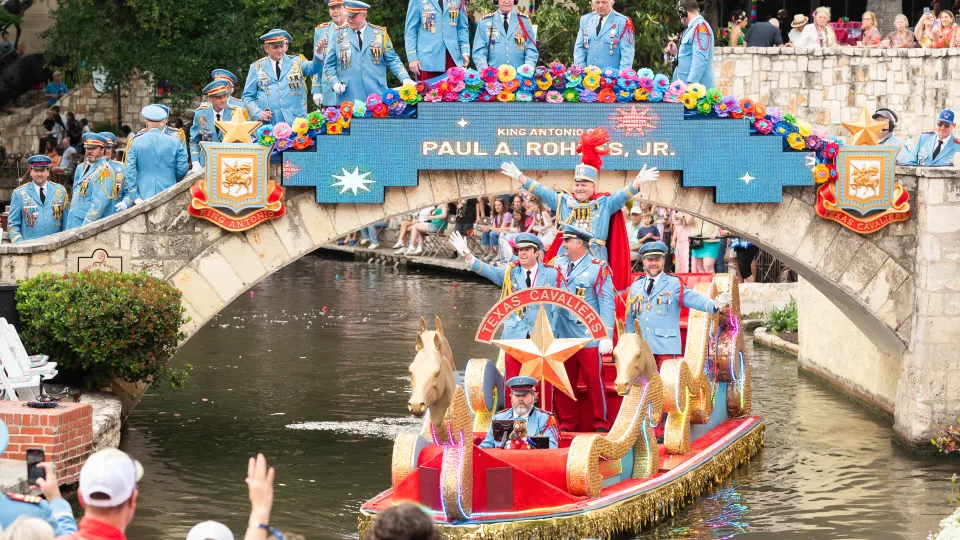 A brightly decorated Texas Cavaliers river barge glides along the San Antonio River beneath a rainbow-fringed bridge, surrounded by participants in light blue uniforms during Fiesta.
