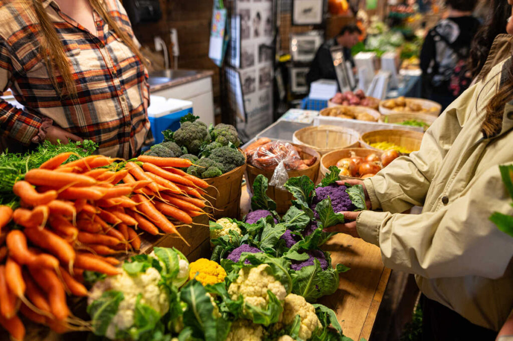 Close-up of fresh carrots, broccoli, cauliflower, onions, and purple cauliflower displayed on a market table, with shoppers nearby.