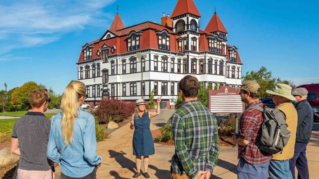 A walking tour group stands outside Lunenburg Academy as a guide gestures toward the striking historic building with its white façade, black trim, and red roof towers.