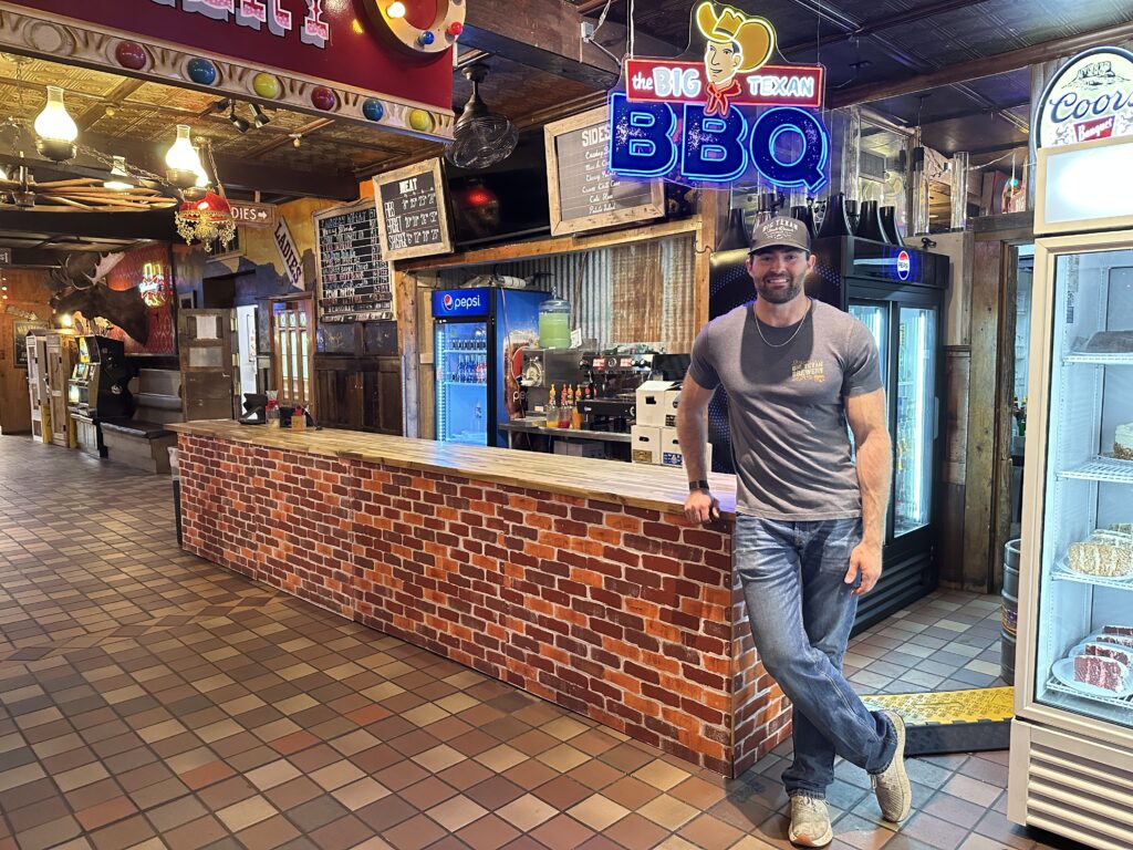 Patrick, third-generation family member of The Big Texan Steak Ranch, standing inside the Amarillo restaurant near a neon Big Texan BBQ sign.