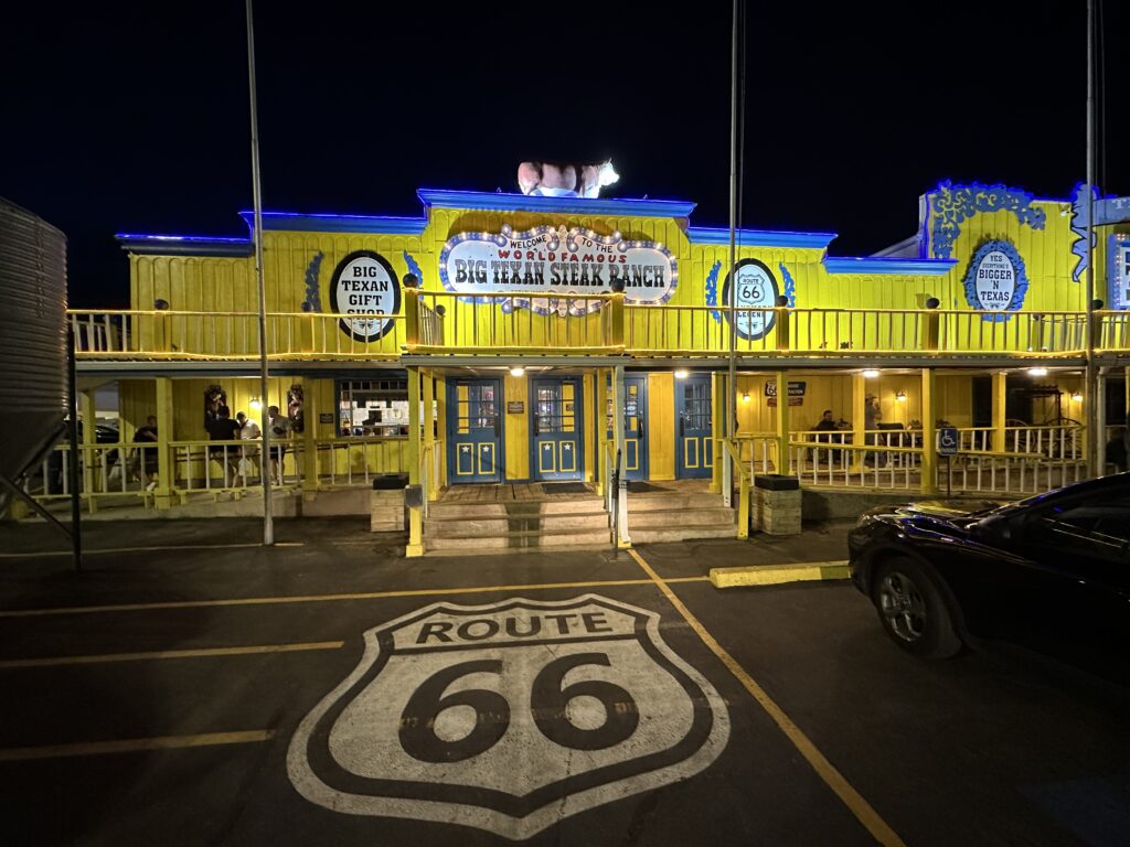 Night exterior of The Big Texan Steak Ranch in Amarillo, Texas, with a Route 66 shield painted in the parking lot.