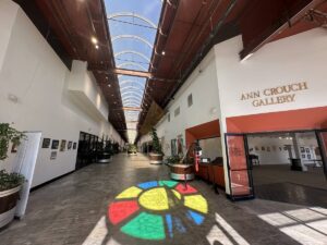 Sunlit corridor inside Arts in the Sunset with the Ann Crouch Gallery entrance, artwork, plants, and colourful light on the floor.