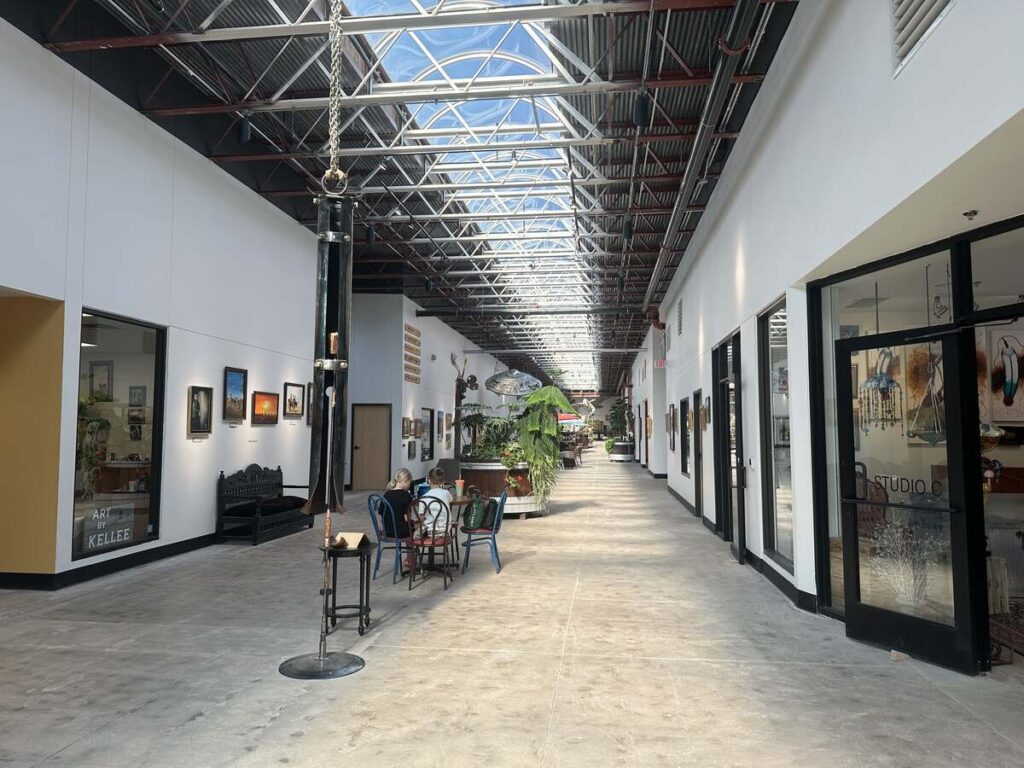 Long skylit corridor inside Arts in the Sunset with gallery walls, studio entrances, plants, and people seated at a small table.