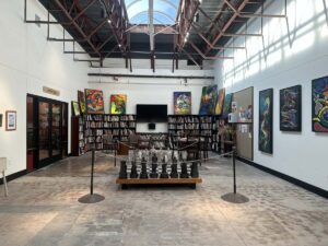 Gallery and library area inside Arts in the Sunset with bookshelves, paintings, a large chess set, and natural light from above.