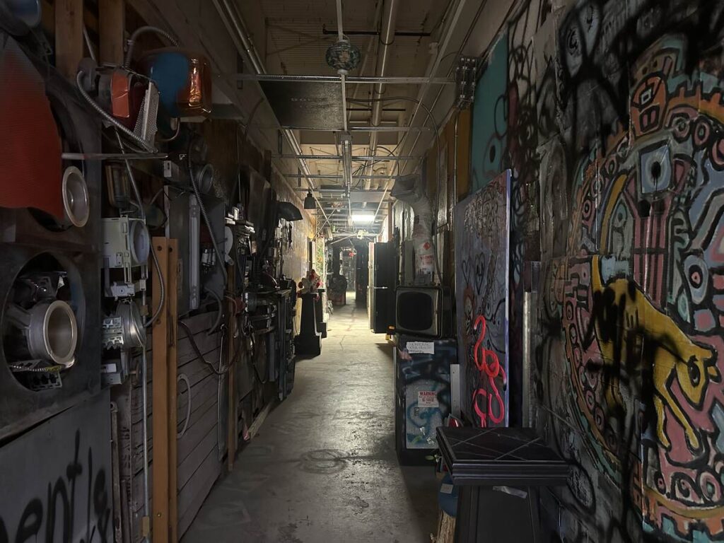 Narrow artist corridor inside Arts in the Sunset with murals, mixed-media pieces, tools, and studio materials along the walls.
