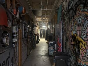 Narrow artist corridor inside Arts in the Sunset with murals, mixed-media pieces, tools, and studio materials along the walls.