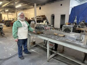 Artist standing in a studio workshop at Arts in the Sunset beside a metal sculpture in progress on a worktable.