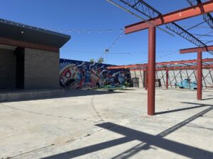 Open outdoor event space at Arts in the Sunset with steel beams, string lights, picnic tables, murals, and a painted car.