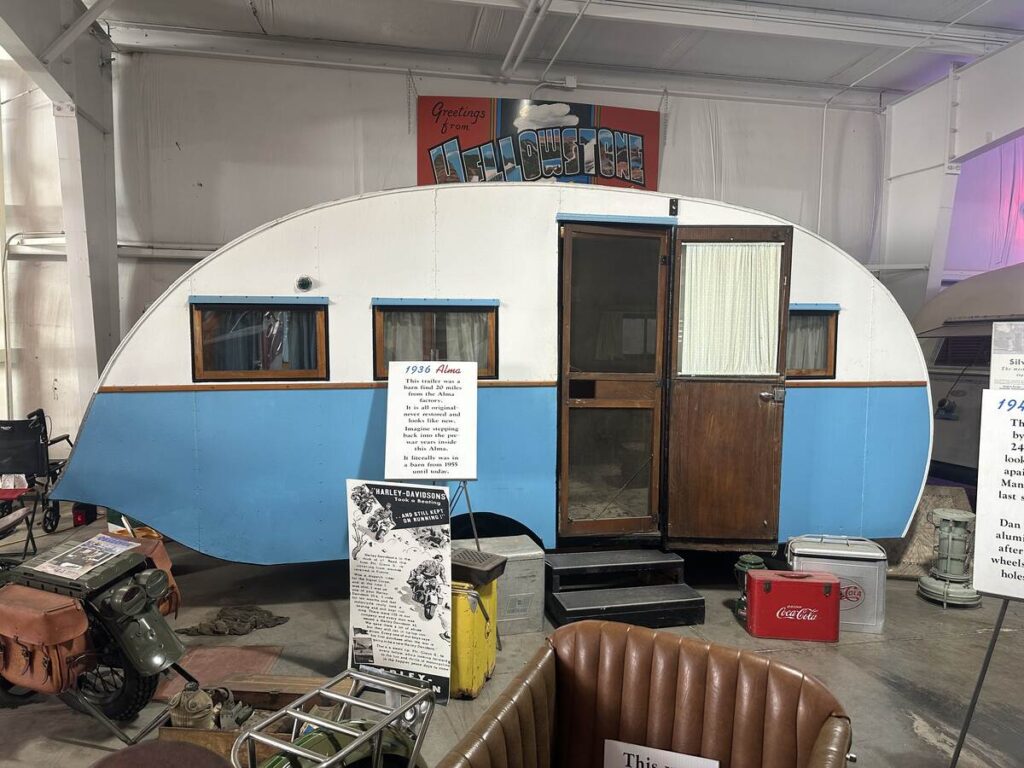 Blue-and-white 1936 Alma trailer displayed at the Jack Sisemore Traveland RV Museum with vintage travel items and signage around it.