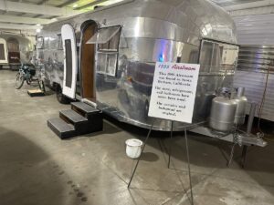 Polished silver 1955 Airstream trailer displayed inside the Jack Sisemore Traveland RV Museum with a sign describing its original interior details.