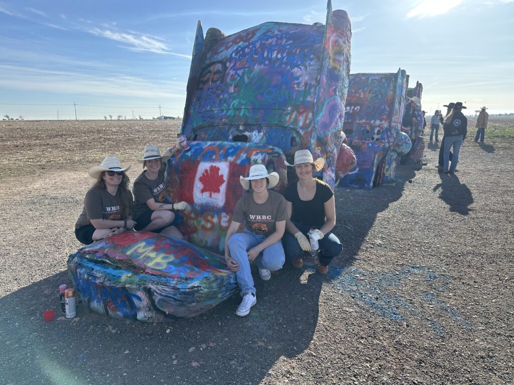 Four women in cowboy hats sitting beside a spray-painted Canadian flag on a Cadillac at Cadillac Ranch in Amarillo, Texas.