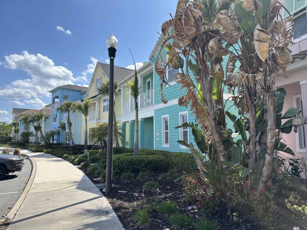 Pastel-coloured resort cottages lined along a sunny walkway with palm trees and tropical landscaping.