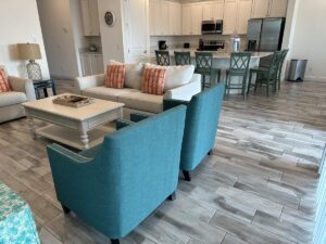 Coastal-style living room with teal chairs, cream sofas, orange pillows, and an open kitchen.