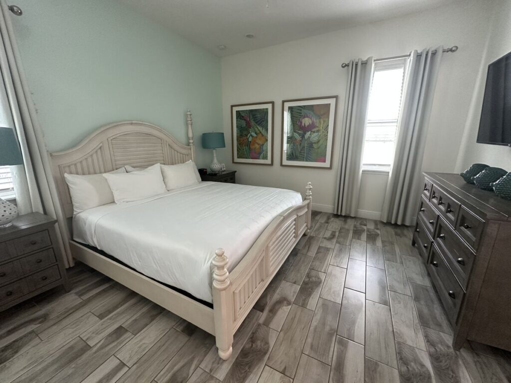 Bright bedroom with white bedding, tropical wall art, grey tile floors, and coastal-style furnishings.
