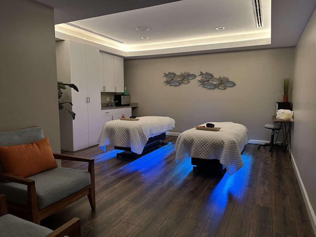 Spa treatment room with two massage tables, soft lighting, wood floors, and fish wall art.