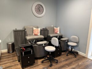 Spa pedicure stations with grey chairs, shell pillows, rolling stools, and neutral coastal decor.