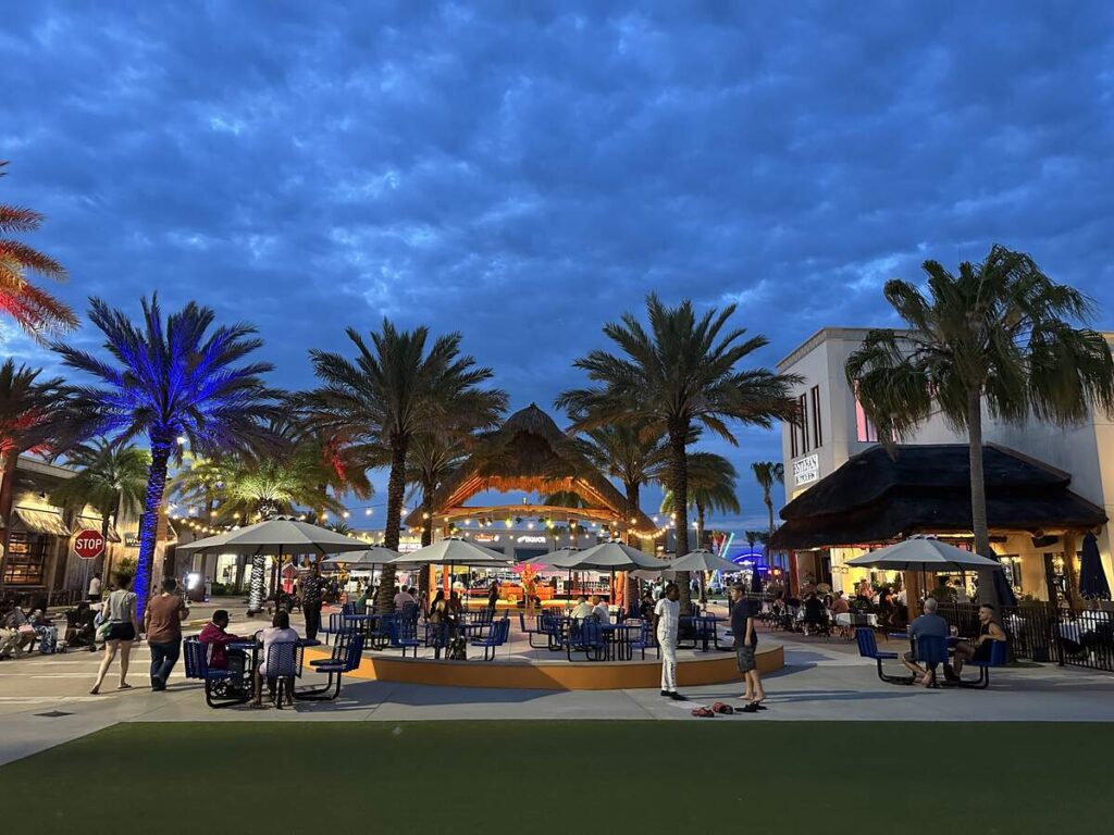 Outdoor entertainment plaza with palm trees, patio seating, string lights, and people gathered at dusk.