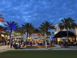 Outdoor entertainment plaza with palm trees, patio seating, string lights, and people gathered at dusk.
