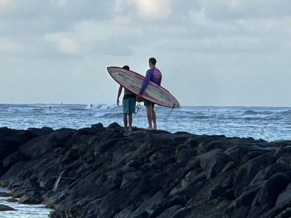 Two surfers standing on dark rocks while looking out toward the waves in Waikīkī.