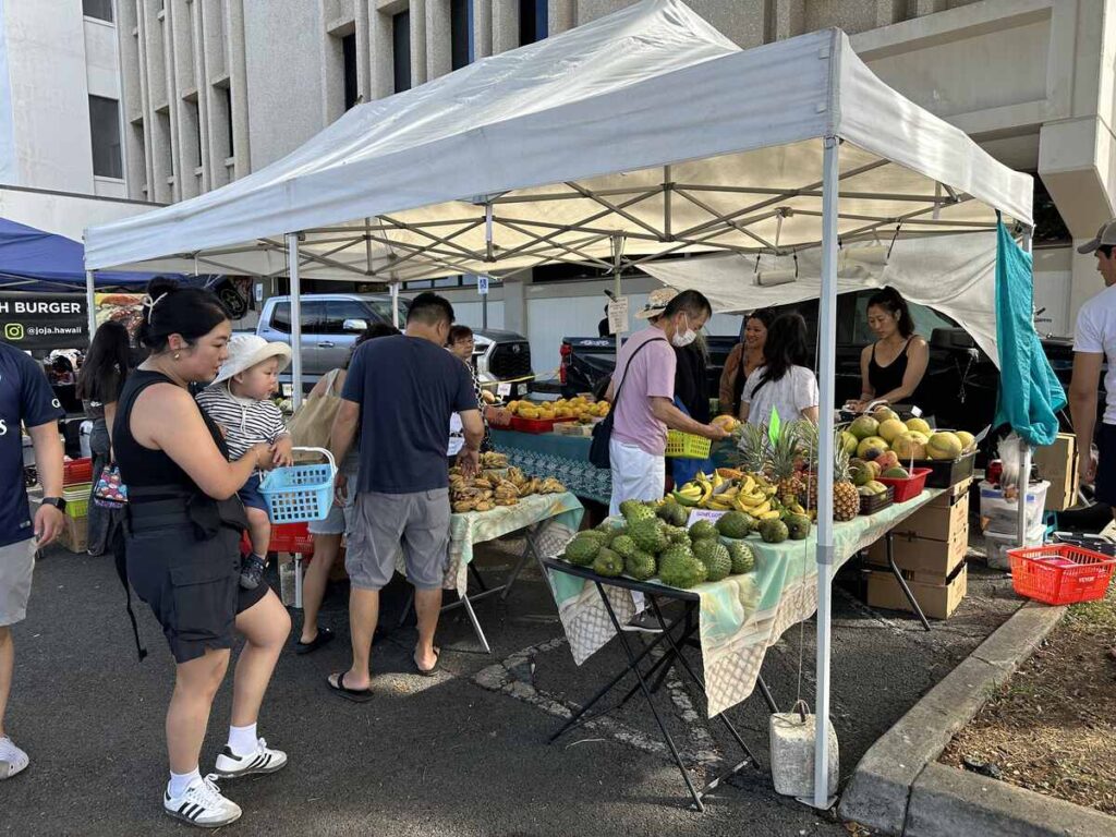 Tropical fruit stand at Kakaʻako Farmers Market with shoppers selecting produce.
