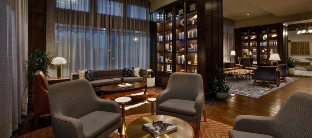 Lounge area inside The Barfield hotel in downtown Amarillo, Texas, with upholstered chairs, wood floors, tall windows, and illuminated bookshelves.