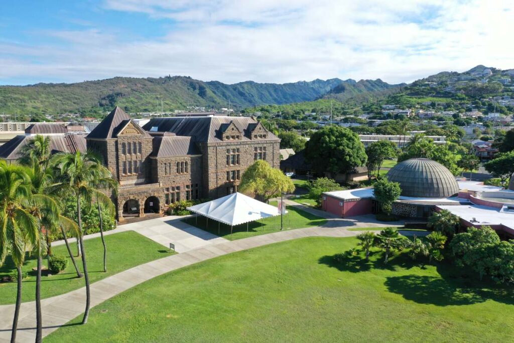 Aerial view of Bishop Museum’s historic campus in Honolulu, with stone buildings, green lawns, palm trees, the planetarium dome, and mountains in the distance.