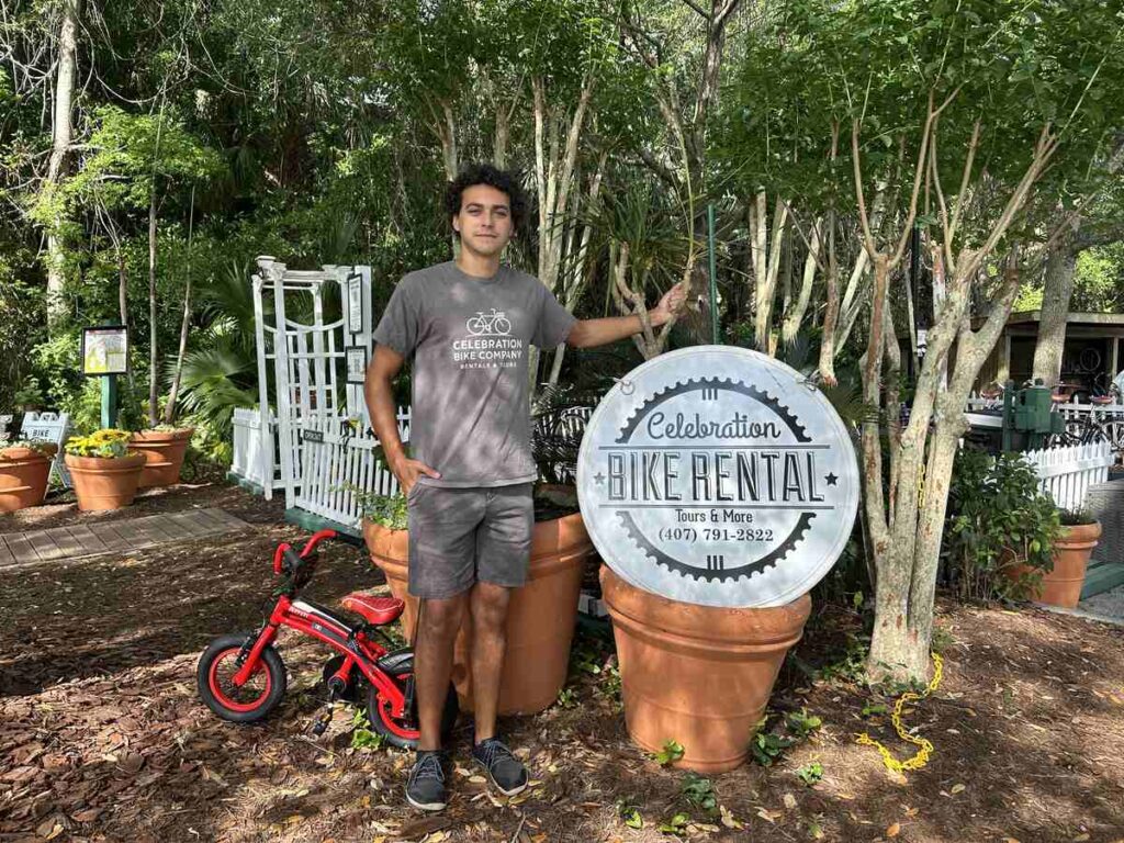 Staff member standing beside the Celebration Bike Rental sign with a small red bicycle nearby.
