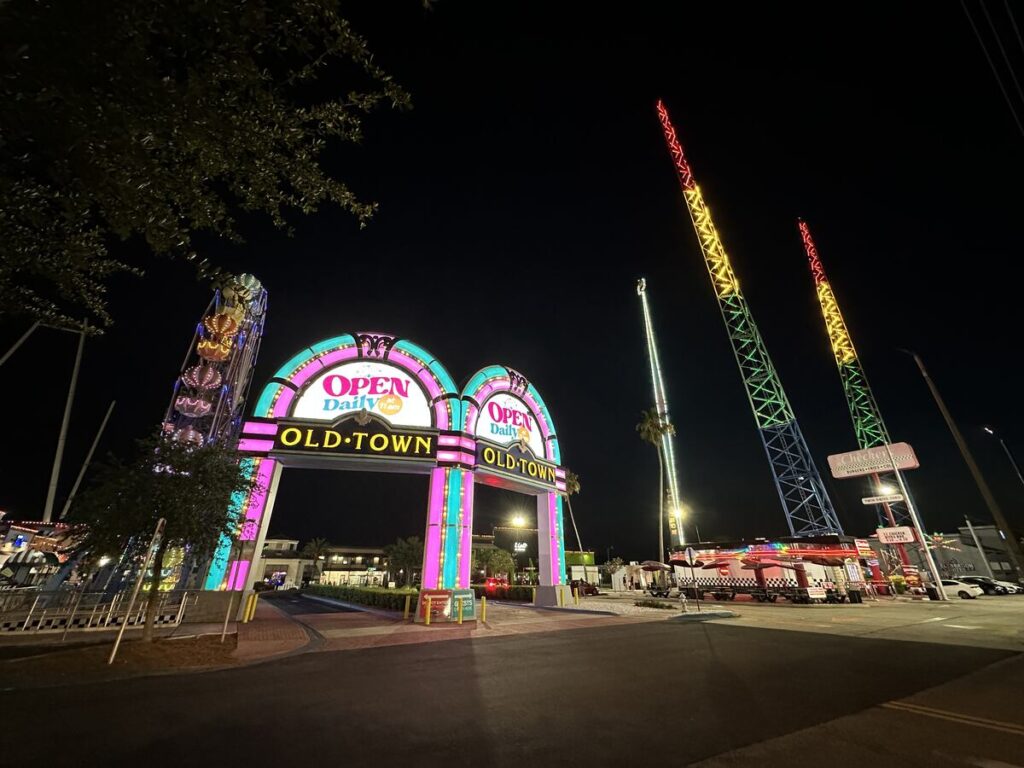 Neon-lit entrance to Old Town Kissimmee at night, with amusement rides and colourful lights in the background.