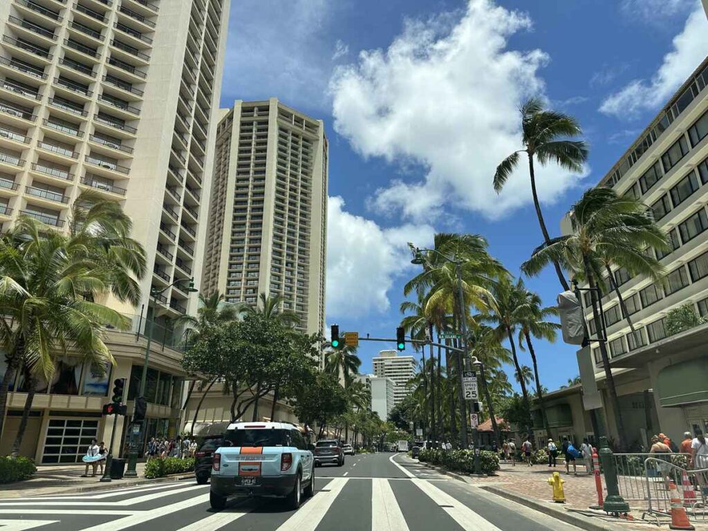 Palm-lined street in Waikīkī with tall hotel buildings, cars, pedestrians, and a bright blue sky.