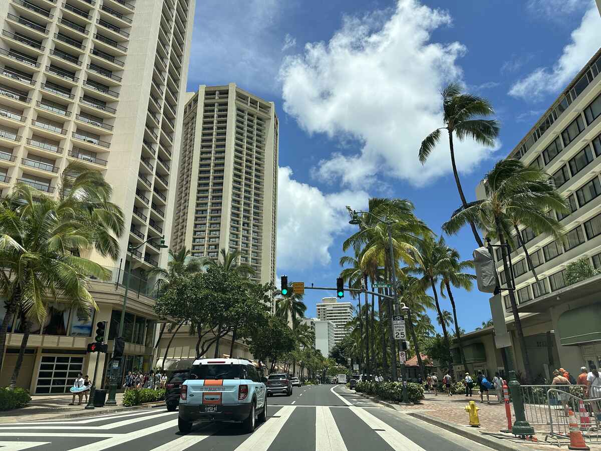 Palm-lined street in Waikīkī with tall hotel buildings, cars, pedestrians, and a bright blue sky.