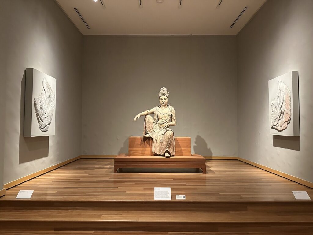 A seated sculpture displayed in a quiet gallery at the Honolulu Museum of Art, with relief artworks on either side.