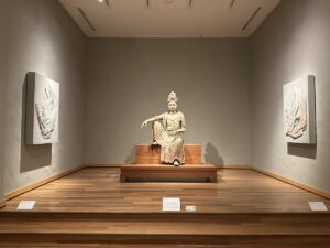 A seated sculpture displayed in a quiet gallery at the Honolulu Museum of Art, with relief artworks on either side.