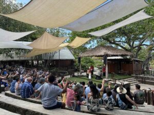 Guests watch an outdoor cultural presentation at the Polynesian Cultural Center, seated beneath shade sails beside traditional-style structures.