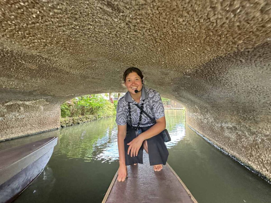 A Polynesian Cultural Center guide crouches on a canoe as it passes beneath a low bridge along the waterway.