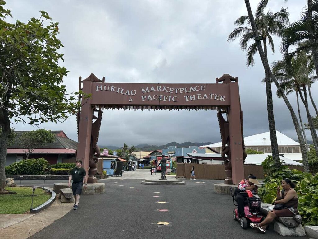 Entrance sign for Hukilau Marketplace and Pacific Theater at the Polynesian Cultural Center, with palm trees and mountains in the background.