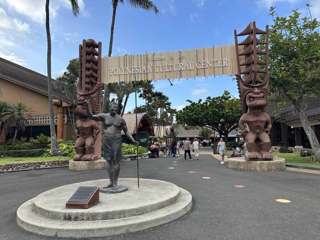 Main entrance to the Polynesian Cultural Center, with large carved figures, signage, and visitors walking through the gateway.