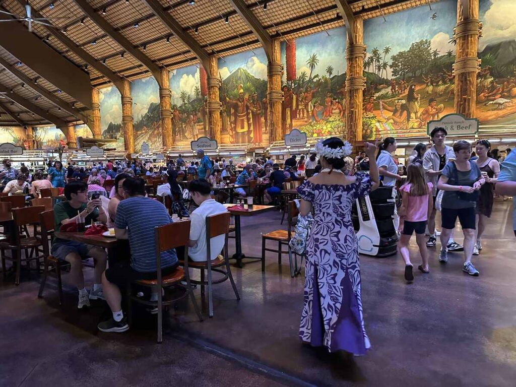 Guests dine inside the Gateway Buffet at the Polynesian Cultural Center, with a large mural and live music in the dining hall.
