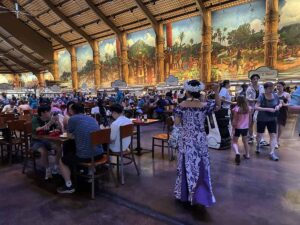 Guests dine inside the Gateway Buffet at the Polynesian Cultural Center, with a large mural and live music in the dining hall.