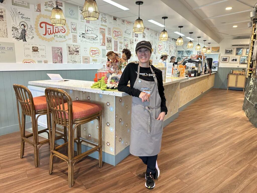 A staff member stands inside Tūtū’s Place by Highway Inn, surrounded by counter seating, woven pendant lights, and a wall filled with restaurant history.