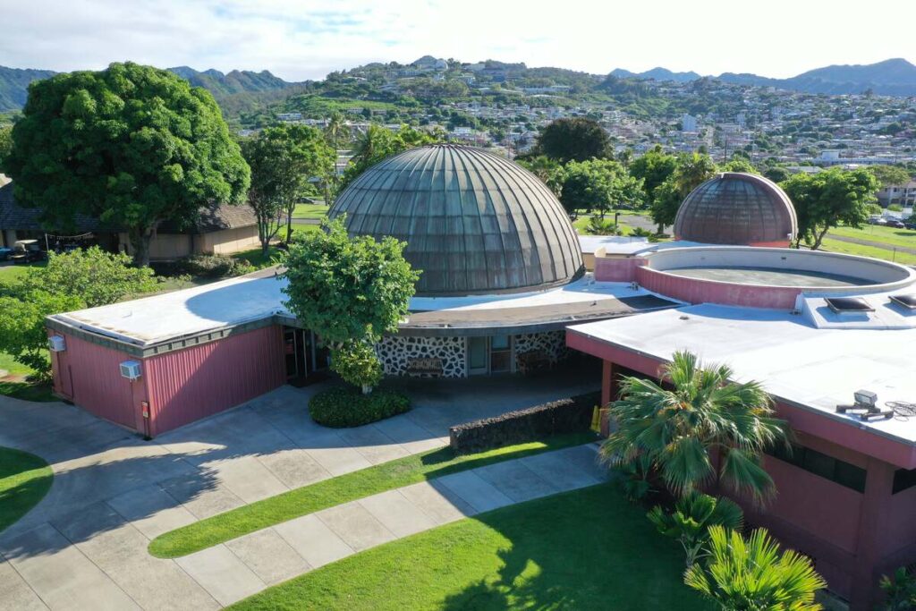 Exterior view of Bishop Museum’s J. Watumull Planetarium, with domed buildings, green lawns, trees, and mountains beyond.