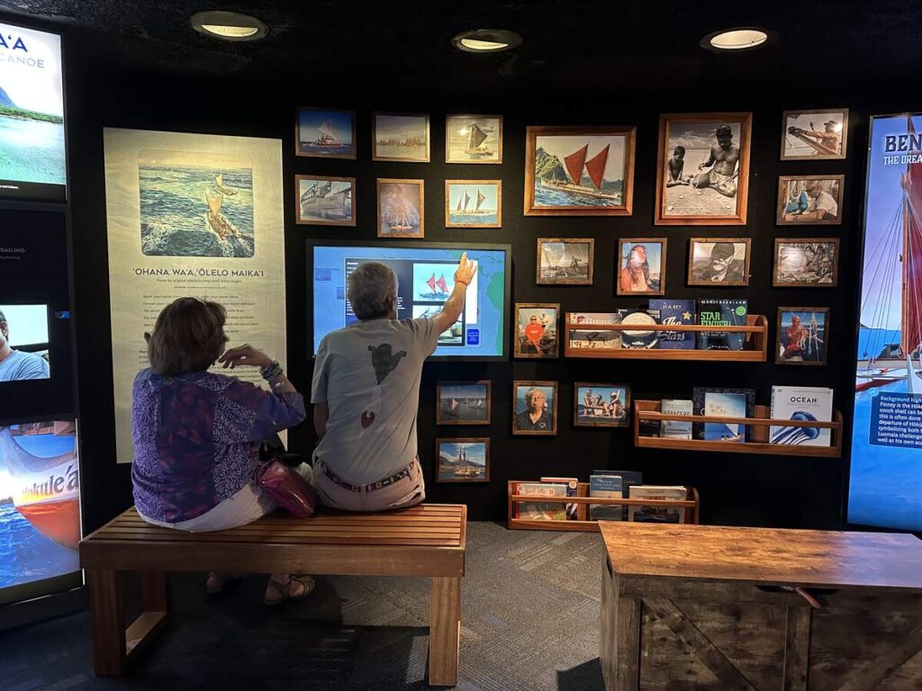 Visitors explore the Wayfinders exhibit at Bishop Museum, surrounded by images, books, and displays about Hawaiian voyaging and navigation.
