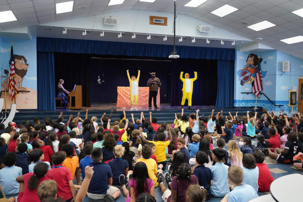 Opera Naples performers present a children’s opera to elementary school students seated in a school auditorium.