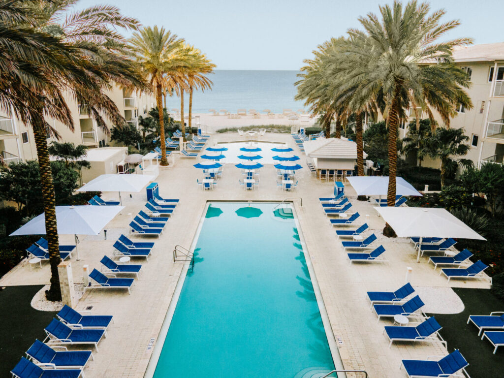 Aerial view of Edgewater Beach Hotel in Naples, Florida, showing the pool courtyard, blue lounge chairs, palm trees, beach, and Gulf of Mexico.