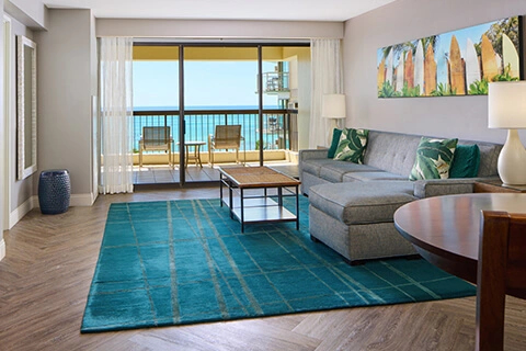 Living room with sofa, turquoise rug, balcony seating, and ocean view at Aston Waikīkī Beach Tower.