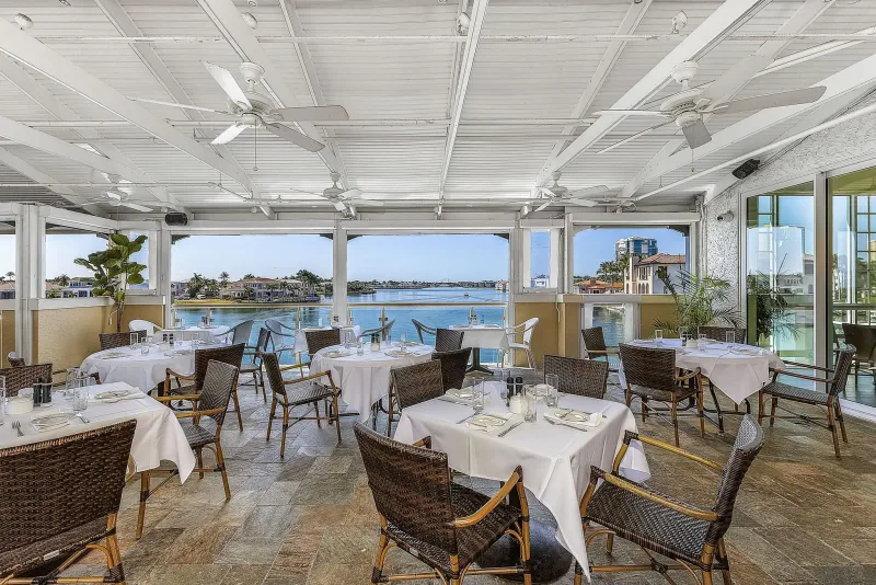 Covered waterfront dining patio at Bayside Seafood Grill & Bar in Naples, Florida, with white tablecloths, wicker chairs, ceiling fans, and views of Venetian Bay.