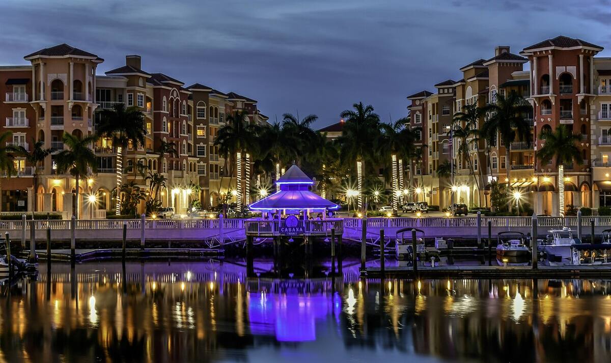 Night view of a waterfront marina in Naples, Florida, with illuminated buildings, palm trees, boats, and reflections on the water.
